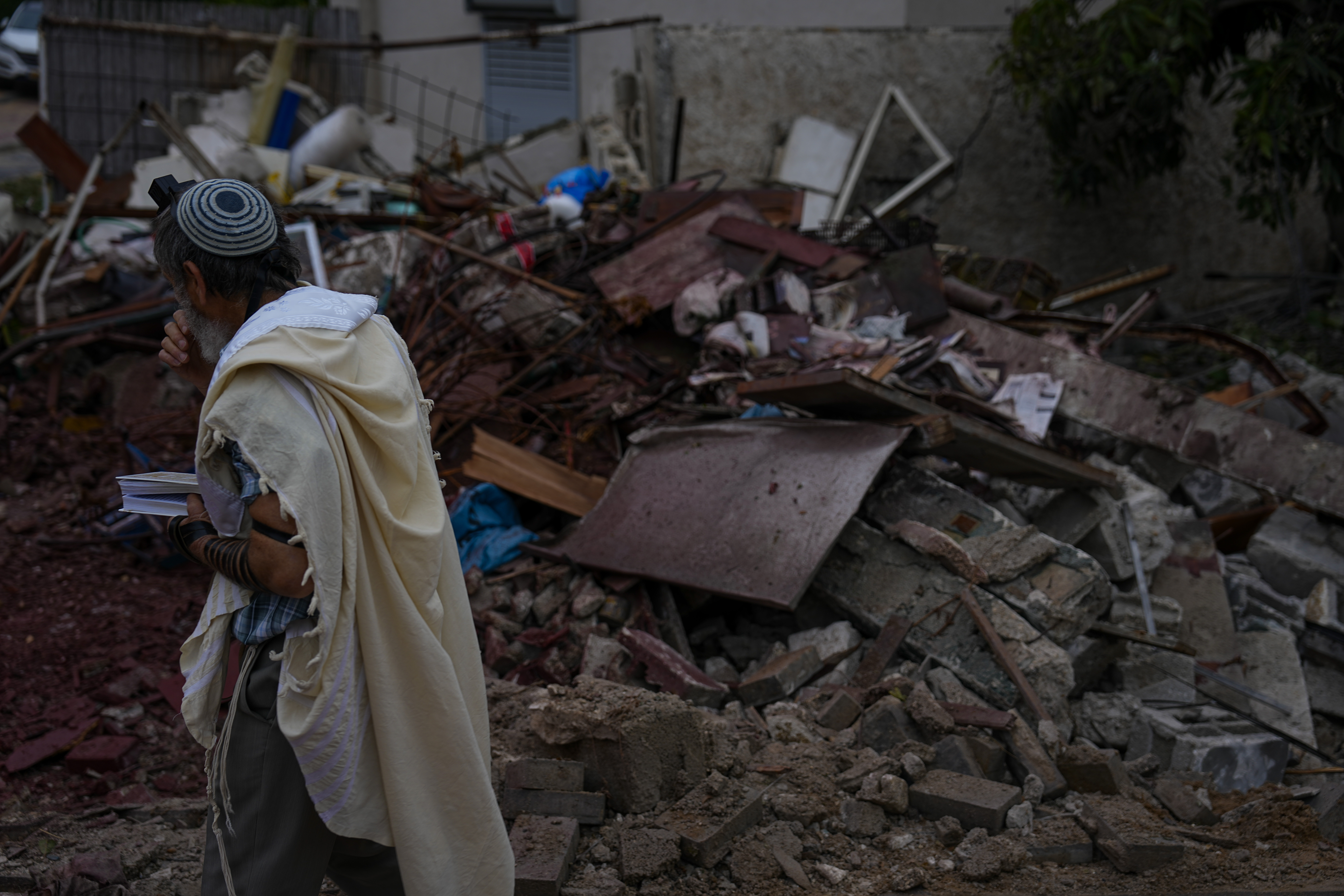 Un hombre cubierto con un manto de oración sostiene un libro sagrado mientras pasa junto a una estructura destruida por un proyectil lanzado en la víspera desde la Franja de Gaza por insurgentes palestinos, en Ashkelon, Israel, el 11 de mayo de 2023. (AP Foto/Ariel Schalit)